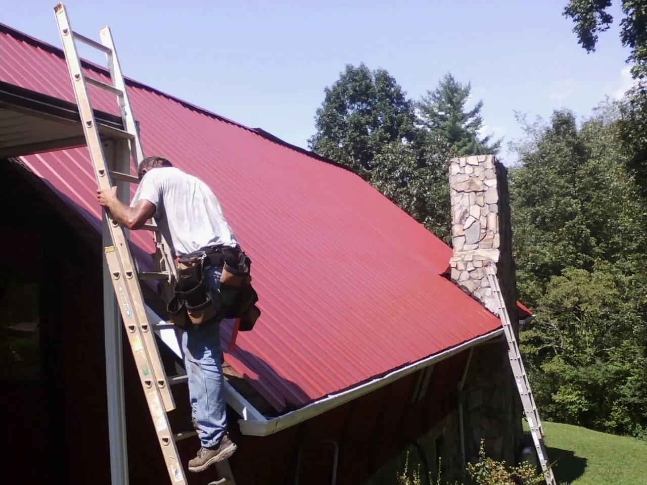 Roofer on a ladder beside a red metal roof and stone chimney — Soffit & Fascia Repair by Roofing Brothers in Stanton